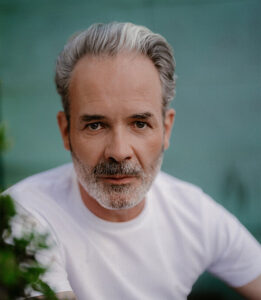 Porträt eines Mannes mit grau melierten Haaren und Bart in weißem T-Shirt.